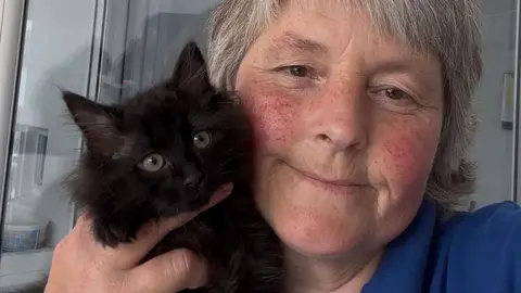 RSPCA Close up of Anne Mitchell holding Buttercup, a fully black fluffy kitten with green eyes. Anne is holding her close to her cheek and she is smiling, has short white hair and rosy cheeks