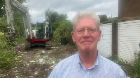 David Hardman looking directly at the camera with a digger in the background. There is rubbish strewn on the floor, grass behind and garages to the right