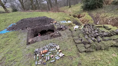 Eddie Goodall A rectangular pit has been dug into the ground in a grassy, wooded area. In the foreground, unearthed glass bottles and metal containers are laid out neatly, showing items that were buried and recently recovered.