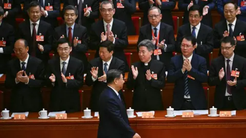 Getty Images Presiden China Xi Jinping, dalam setelan hitam, berjalan melewati para pemimpin partai lainnya yang berdiri dan bertepuk tangan saat ia tiba untuk upacara pembukaan Konferensi Konsultatif Politik Rakyat Tiongkok (CPPCC) di Aula Besar Rakyat di Beijing pada 4 Maret 2025. 