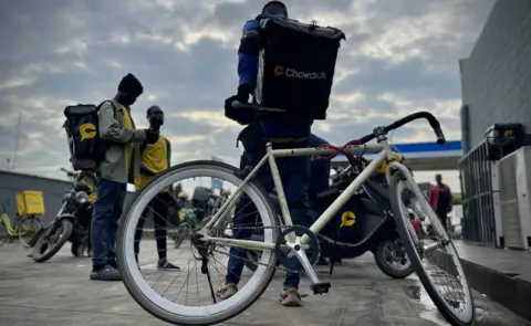 Gift Ufuoma/BBC A group of men standing with motorcycles parked around. One man is sitting on a bicycle.
