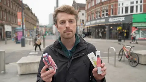 Man holding two disposable vape boxes in both hands stares at camera