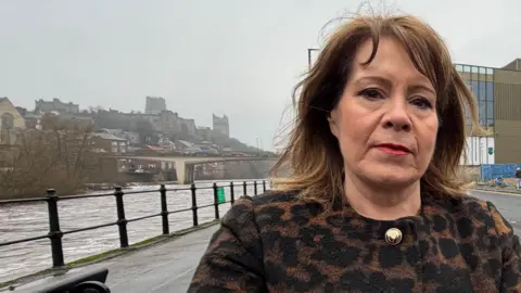 MP Mary Kelly Foy standing across the road from the almost finished Milburngate development. Foy is looking at the camera with a serious expression. She has shoulder-length brown hair and is wearing a leopard print-type coat with golden buttons. To the left is the river Wear and to her right is the development. It is made of a main building with window walls that looks finished. In front of it is heras fencing around the perimeter.