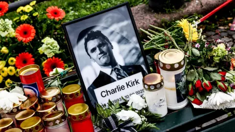 EPA A black and white framed photo of Charlie Kirk, wearing a suit, is in the centre of a vigil. Next to the photo, there are candles and red and yellow flowers. 