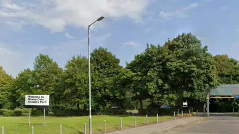 Google Maps Green trees, green grass and a blue sky. On a roadside verge is a white sign reading: 'Welcome to Moses Gate Country Park'.