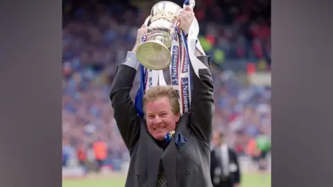 A man standing at the edge of a football pitch holds a large trophy high above his head as he celebrates. He has light hair and wears a grey suit. The trophy has long strips of ribbon attached to it with the Nationwide Building Society logo printed on them. Crowds in the stands are blurred behind him. 