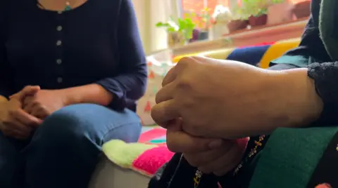 Two people sitting down in a room, showing their hands. One is wearing jeans, with a blue top, the other has their hands clenched. 