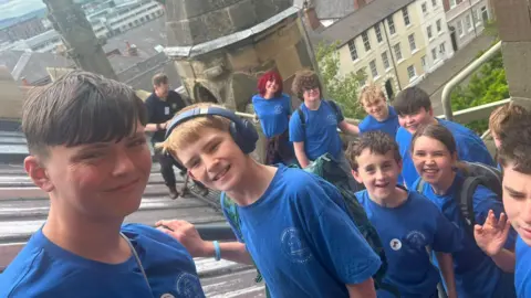 Sam Martin A group of young people are standing outside, on a church tower. They are smiling for the photograph. Most of the group are wearing blue t-shirts.