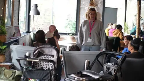 Jamie Niblock In the foreground are two baby prams. Four women are sitting on the sofas with two holding babies. Another woman - with blonde shoulder-length hair is standing talking to them. 