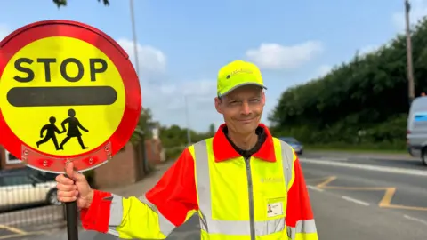 BBC News A lollipop man wearing his high vis yellow and orange uniform holding his lollipop stick and smiling at the camera, next to the road where he works