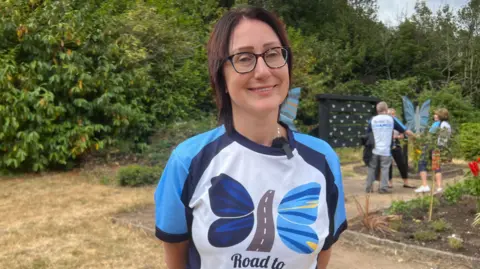 A woman with a white top looking into the camera and smiling. She has got a butterfly on her top and it has a road in the middle of it.