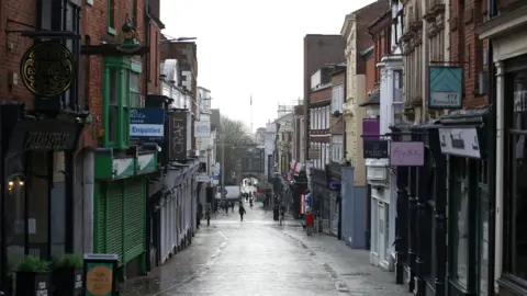 A view of a shopping street looking downhill. There is a row of shops on either side of the cobbled street. The shops are closed with shutters down on a few of them.