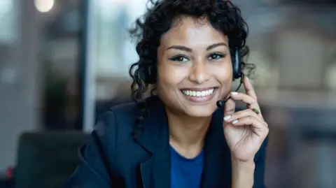 A woman wearing a dark blue blazer and blue t-shirt and a headset smiles towards the camera, seemingly during a call