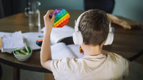 Picture is by Getty, and posed by a child model. The child, who is about 9 or 10, is sitting facing away from the camera at a table, wearing white headphones and writing in a workbook. In his left hand is a rainbow fidget toy.