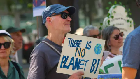Joao Santos/Local Democracy Reporting Service A man at a protest who looks to the right. He has a blue cap, sunglasses and is wearing a dark blue T-shirt. He is holding a cardboard sign that reads there is no planet B. Other protesters 