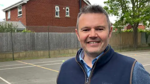 A man in a blue jumper and a navy gilet stands in a playground smiling at the camera. 