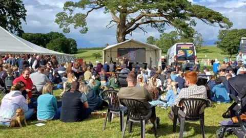 A sunny day and lots of people sitting watching a big screen watching a football match on a big screen 