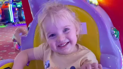 PA Isabella Jonas-Wheildon, a young blonde toddler wearing a yellow t-shirt smiling at the camera. She is sitting on a yellow and purple chair at an arcade.
