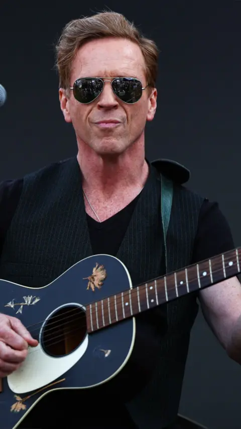 Damian Lewis wearing sunglasses and black t-shirt holding guitar