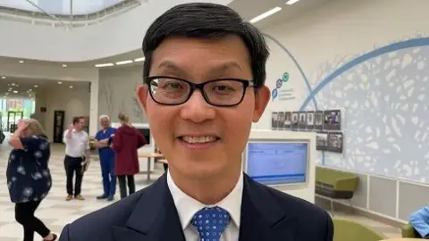 Nadia Gyane/BBC Steven Tsui with short dark hair and black-framed glasses looking at the camera and wearing a dark blue jacket with white shirt and blue tie with lighter blue spots. There are people behind him walking across a large foyer area of a hospital. There is a blue pattern on the wall to Ms Tsui's left. Glass doors can be seen in the background.