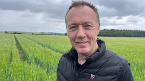 Neil White, with short, dark hair and wearing a black bodywarmer and tee-shirt, looks to the camera in front of his crop of spring barley.