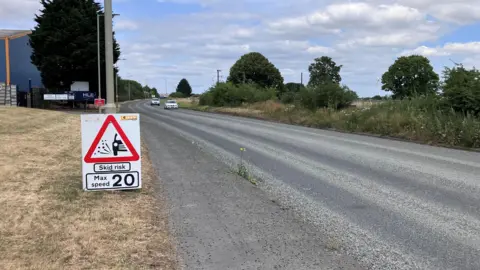 A newly resurfaced road with no road markings. On the left is a path, grass and an entrance to an industrial site in the distance. There is a skid risk sign against a lamppost on the grass. It says the maximum speed is 20mph. Two cars can be seen travelling towards the camera in the distance.