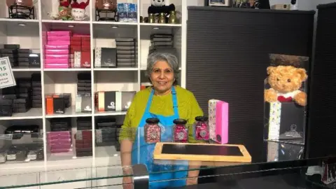 BBC A woman standing behind a chocolate shop counter. She is wearing a green jumper and blue apron. She is smiling at the camera and has thick, grey hair. Boxes of chocolate are in an open, white cupboard behind her. 