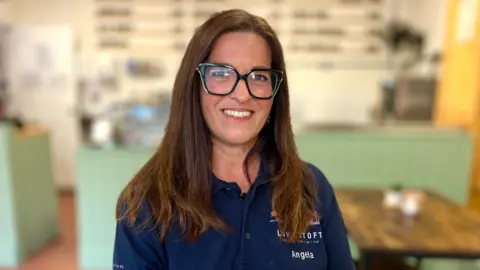 Martin Giles/BBC Angela Grey is sitting in a coffee shop and you can see the counter behind her and the menu on the walls. She is wearing a blue polo shirt with a Lowestoft logo on it. Angela is wearing glasses and is smiling at the camera 