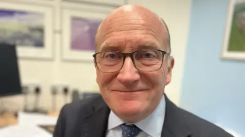 Nick Hulme, a balding man wearing a grey suit jacket, white shirt, blue spotted tie and glasses smiles at the camera. The blurred background appears to show a desk and monitor.