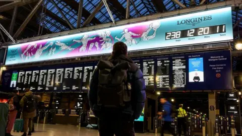 A man with a backpack standing in a railway station, looking up a the large departure boards and the countdown clock of the Commonwealth Games - a blue and purple designed illustration of athletes playing sport, with a clock ticking down to the Games starting. 