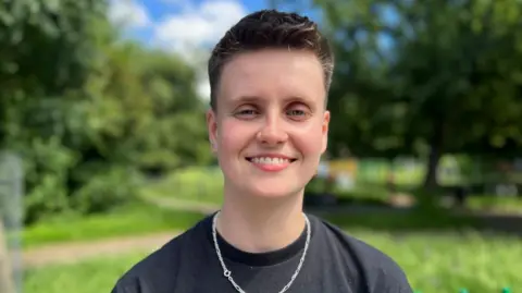A woman smiles at the camera with trees and grass behind. She's got short cropped dark-blonde hair and is wearing a dark grey t-shirt with a black seam, and a silver chain around her neck. 