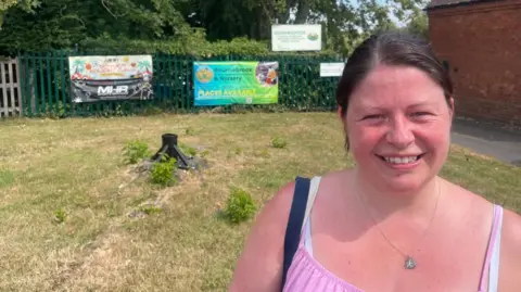 A woman stands to the right of the photo smiling at the camera. She has brown hair in a ponytail and is wearing a pink top with straps. Behind her is green railings with two signs on for Bournebrook Church of England Primary School,