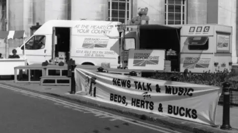 A black and white picture of a BBC van and truck on a street in Luton. They are part of an outside broadcast. A banner advertises the radio station for being a good place for music, news and talk shows.