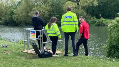 Sam Read/BBC Two police officers are stood on a river bank and speaking to two men. One of the men has a red shirt with sleeves rolled up. The other man has his back to the camera and is looking out to the river. A small drone is parked on the grass.