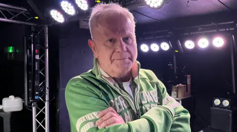 Tom Jackson/BBC Steve Jason is standing on a small stage which has stage lighting on its ceiling and walls. Jason has short white hair and he is wearing a white T-shirt, and a green jacket which has white writing and stripes. 
