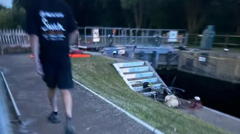 Wendy Hermon A slightly blurred photograph of a canal lock. On the left, a person wearing a black T-shirt walks away from the camera along a paved towpath. On the right is the lock with a short flight of steps and some equipment at the bottom.