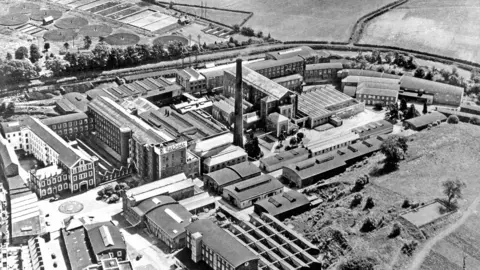 Irish Linen Centre & Lisburn Museum A black and white aerial photo of the Hilden Mill site. The mill itself stands tall in the centre of the image, with multiple buildings surrounding it. Fields and trees are on the outskirts of the photo.