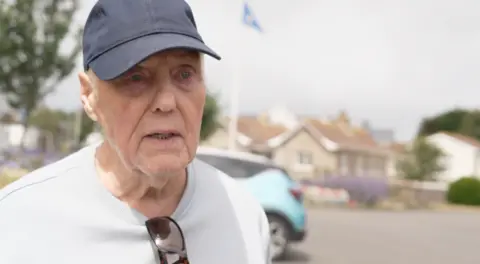 An older man is pictured in a car park, with a blue cap and white tshirt.