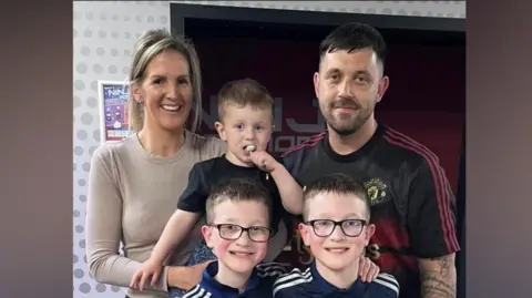 Family photo Hudson with his parents and twin brothers. His mother Natalie holds Hudson and father Damian stands to their left. His identical twin brothers stand in front of them, wearing matching navy football tops. Hudson has a lollipop in his mouth.