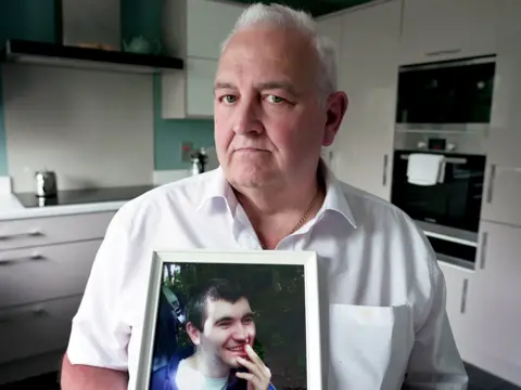BBC Glynn Brown stands in his white kitchen holding a framed photo of his disabled son Aaron. He looks into the camera with a serious expression and the photo of Aaron features him smiling.