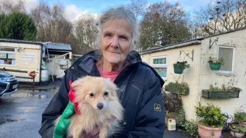 Marty pictured looking into the camera and smiling slightly. She's wearing a long navy waterproof coat with a pink top on underneath, and has her long grey hair tied up out of her face. She's holding a small light brown fluffy dog, possibly a Pomeranian cross. The dog is looking towards the camera, and has a green and red Christmas elf hat on its head. They're stood outside the rescue centre, which is made up of a cluster of single-storey cream buildings. 
