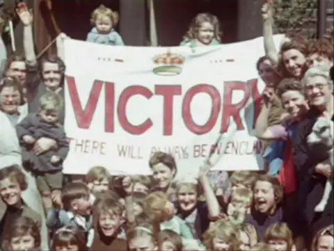 A crowd of people celebrating Victory Europe Day in 1945. The crowd is made of a mix of young and old. They stand in front of a large white banner that reads, VICTORY - THERE WILL ALWAYS BE AN ENGLAND.