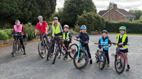 A group of children and adults on bikes are in an empty car park. The are wearing cycling helmets and high visability vests and smiling at the camera. There is greenery behind them and a brick built house in the distance.