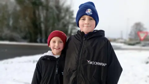 Two children, a boy and girl, in winter clothes standing outside in the snow in front of a street.