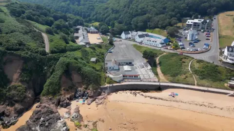 BBC A drone photo of Greve De Lecq beach. At the bottom of the photo is the beach with browny-white sand, and on the left the sand turns into brown cliffs with green grass and hedges on top of them. There is also a car park and conrete.