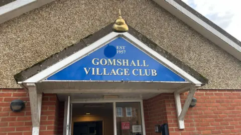 Clare Cowan/BBC The entrance to Gomshall Village Sports & Social Club. Above the door is a sig with a blue background and gold writing saying "Gomshall Village Club"