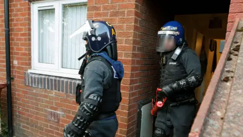 Greater Manchester Police Greater Manchester Police officers taking part in drugs raids at a house in Gorton. Officers are carrying tools to break down doors in the strikes and wearing riot gear.