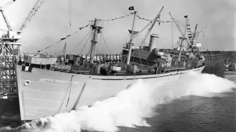 A black and white photograph shows the James Egan Layne Liberty Ship causing a wake as the boat is launched into the water for the first time in a dockyard 