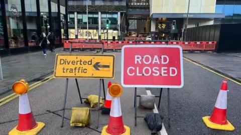 BBC A road is blocked off by cones, orange fencing and a sign which says "diverted traffic" and another which says "road closed"