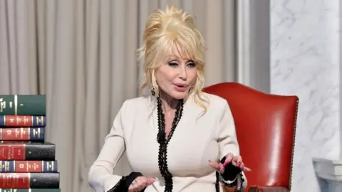 Shannon Finney/Getty Images Dolly Parton, wearing a black and white blazer, sits on a red leather chair and speaks to a crowd. There is a pile of old-style books next to her.